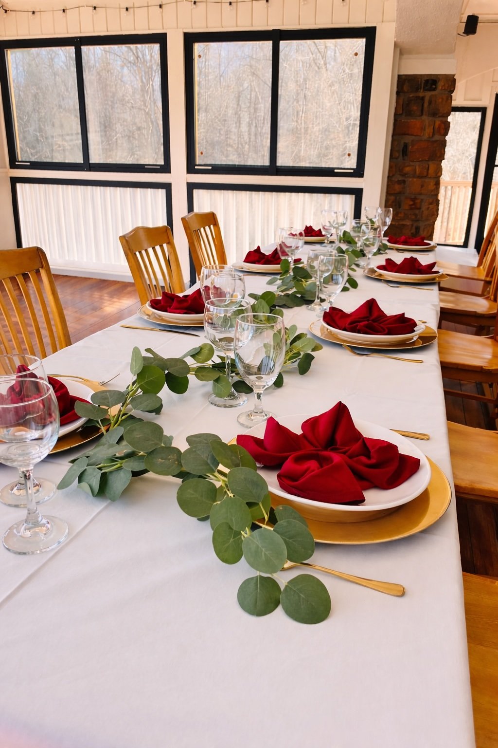 A dining table set for a meal with white tablecloth, gold-rimmed plates, red cloth napkins folded on plates, wine glasses, and green leafy garland centerpieces, in a bright room with large windows and wooden chairs.
