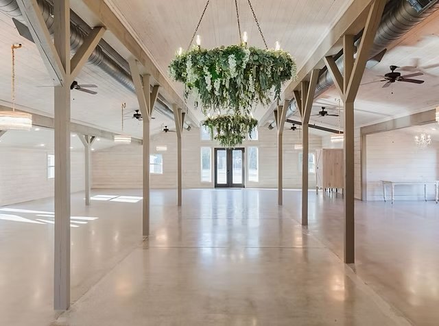 Empty wedding venue with wooden beams, chandelier with greenery, ceiling fans, large windows, and light-colored wooden walls.