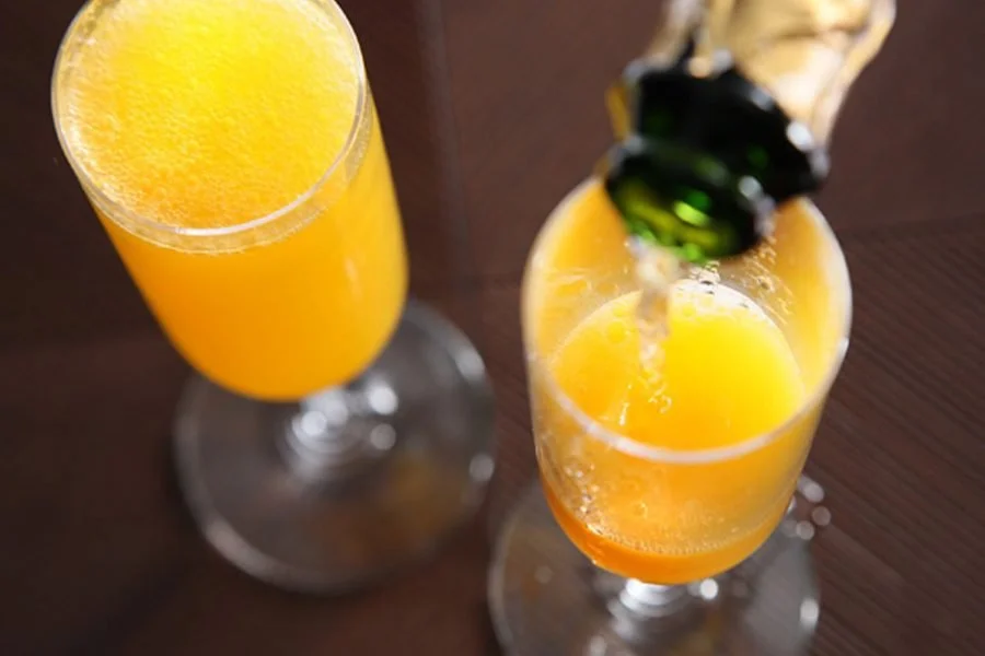 Two champagne flutes filled with orange juice, one being poured from a green bottle, on a dark wooden surface.
