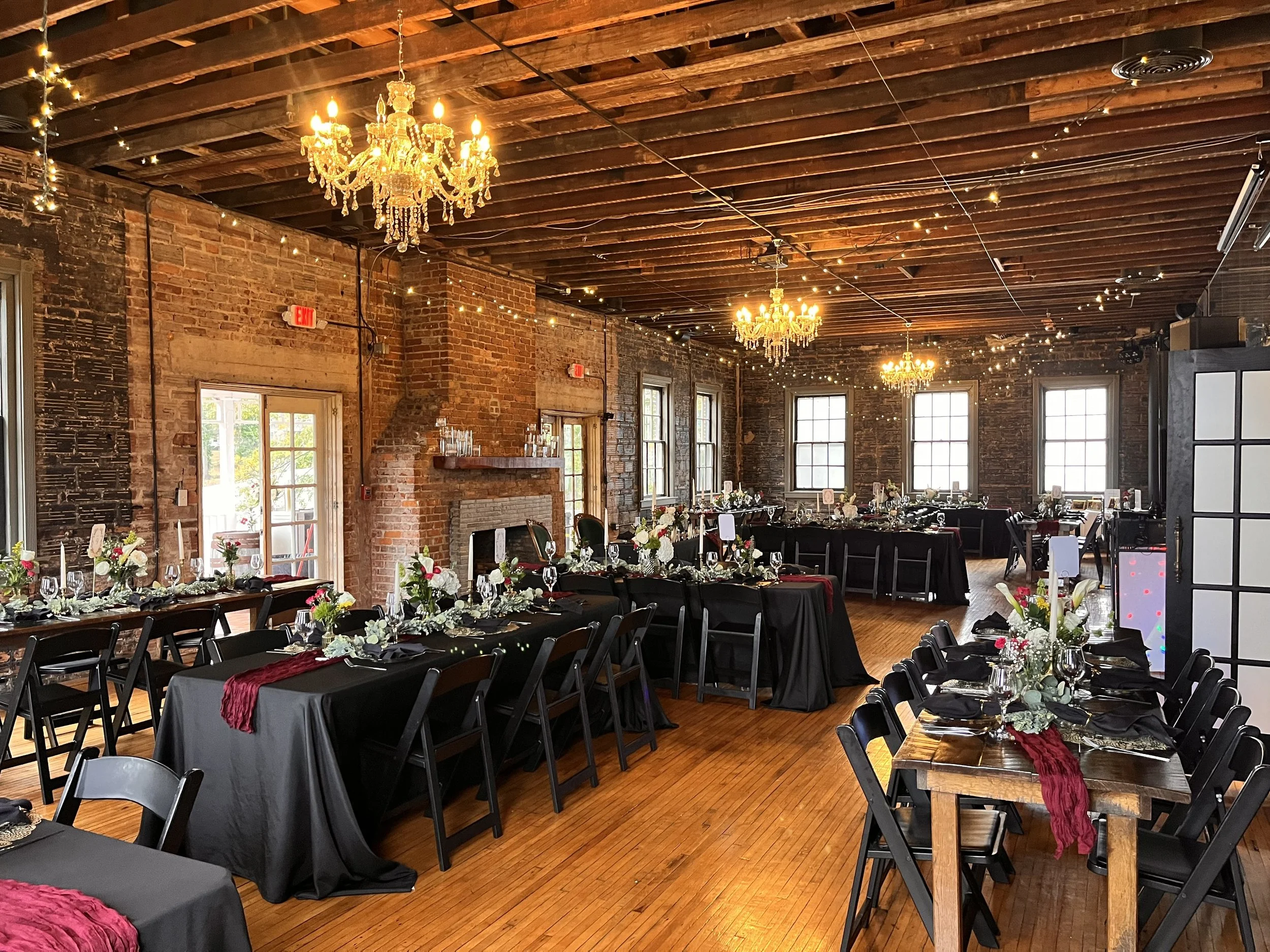 Elegant banquet hall decorated for an event with black tablecloths, floral centerpieces, and chandeliers, featuring exposed brick walls and large windows.