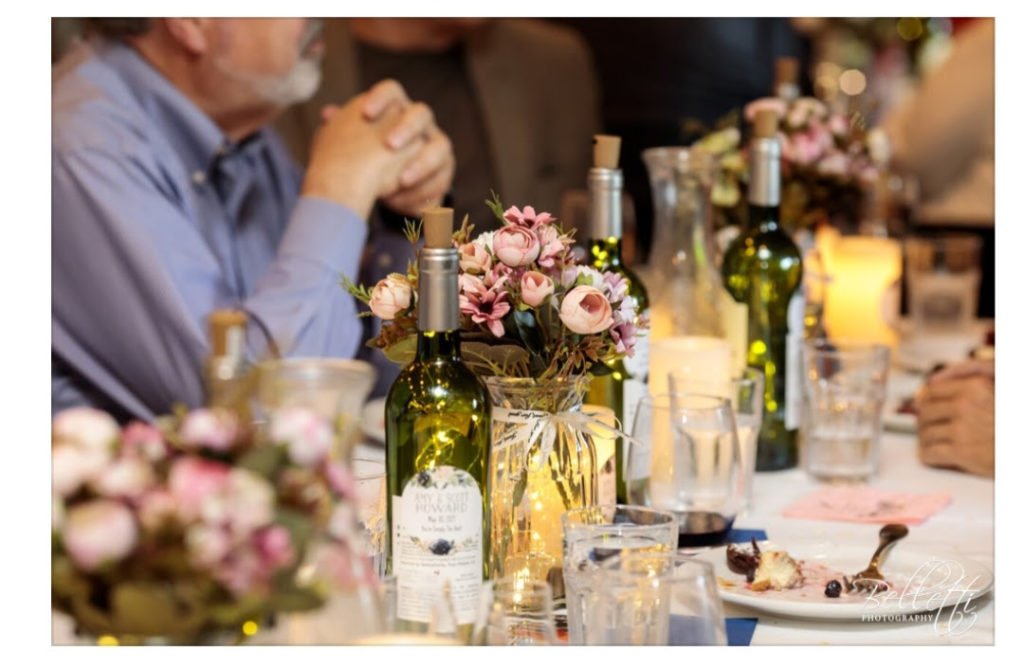 A wedding or celebration table decorated with pink flower arrangements, wine bottles, and wine glasses, with people sitting around.