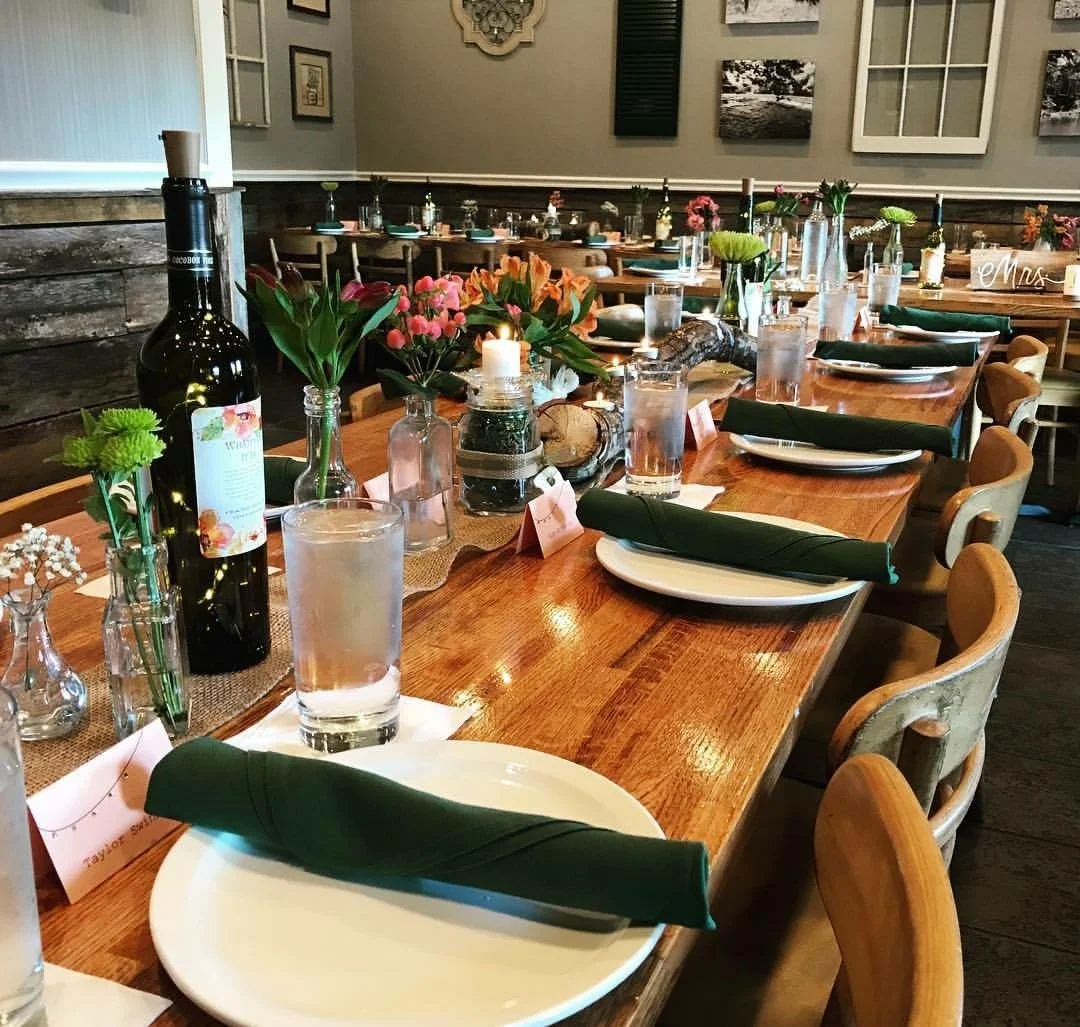 Decorated dining table with flowers, candles, wine, water glasses, plates with green napkins, and place cards, set for a celebration or event in a rustic-themed restaurant or venue.