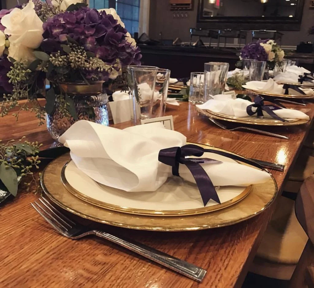 A formal dinner table setting with white napkins tied with black ribbons on gold-rimmed plates, clear drinking glasses, and floral centerpieces with purple and white flowers.