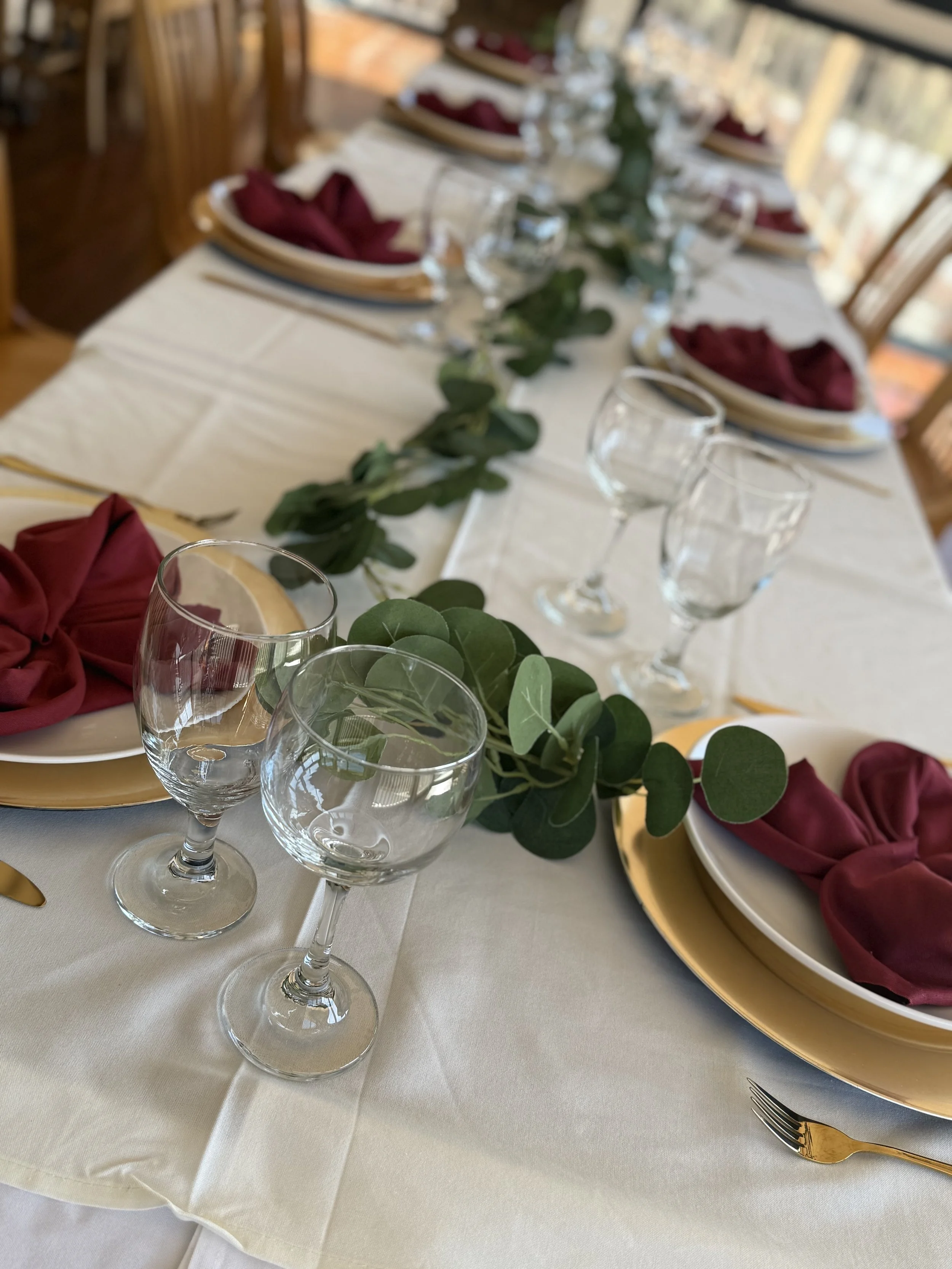 Elegant dining table set for a formal meal with white tablecloth, gold-rimmed plates with burgundy napkins, clear wine glasses, and a eucalyptus garland centerpiece.