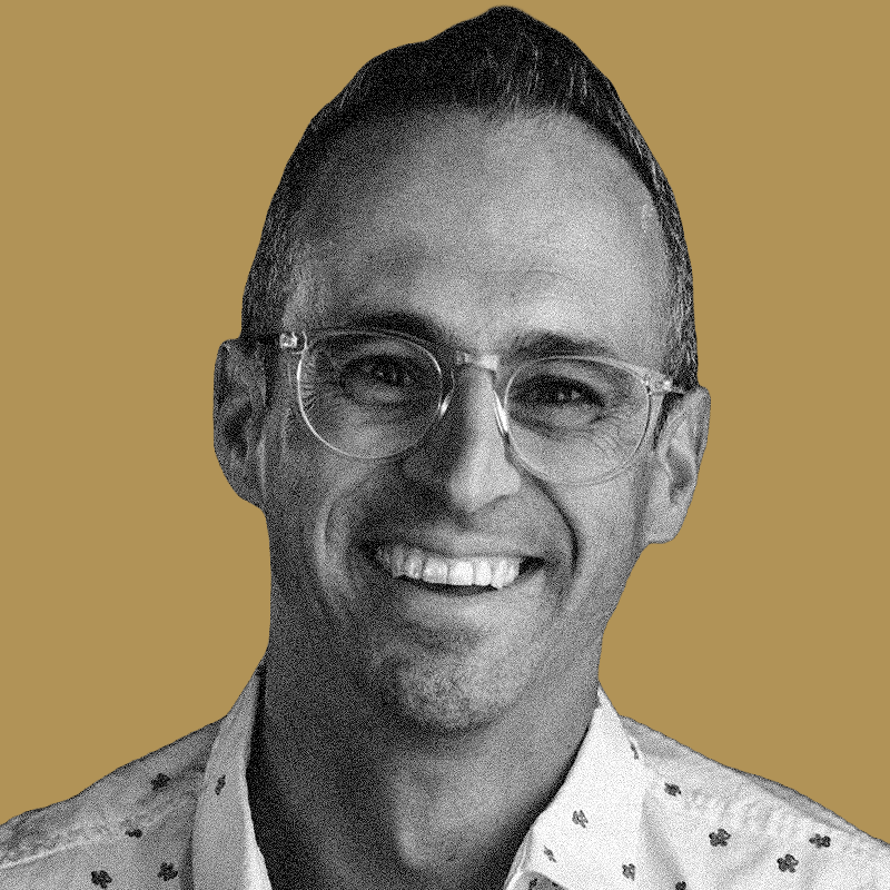 A smiling man with short dark hair, glasses, and wearing a patterned shirt standing against a plain color background.