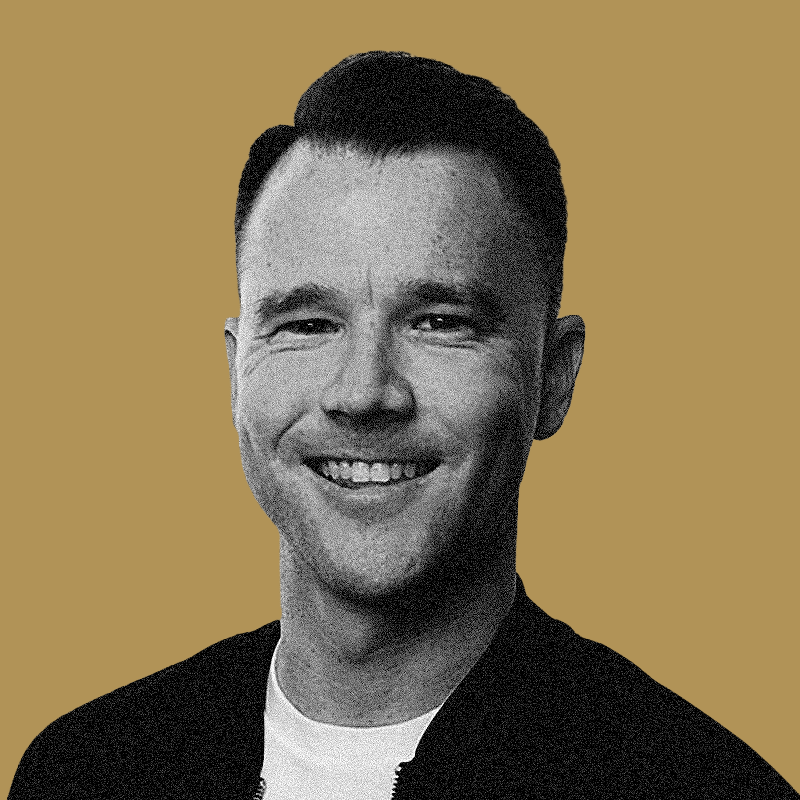 Black and white portrait of a man smiling, wearing a jacket and a white shirt, with a plain beige background.