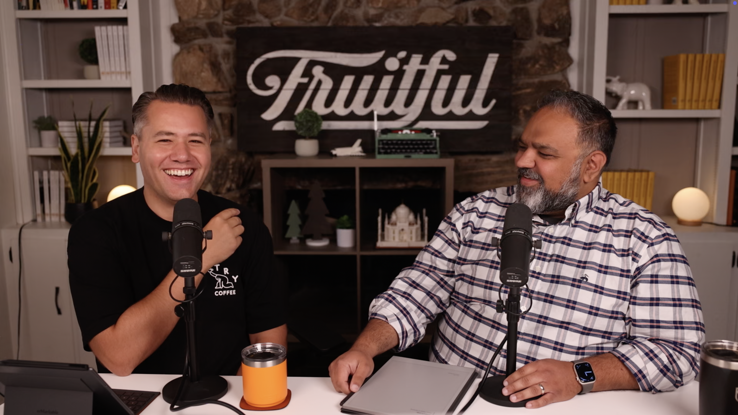 Two men sitting at a desk with microphones, one smiling and the other looking at him, with a decorative background that says 'Faithful' and contains books, plants, and ornaments.