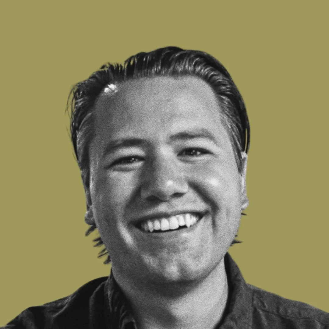 Black-and-white photo of a smiling man with short, slicked-back hair, wearing a dark shirt, against a plain, muted background.