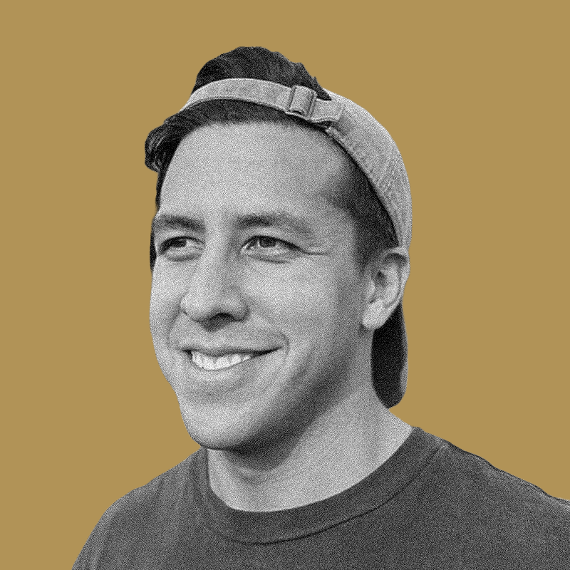 Black and white photo of a smiling young man wearing a backwards baseball cap and a dark t-shirt, with a plain tan background.