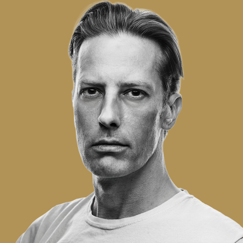 Black and white portrait of a man with slicked-back hair wearing a light-colored T-shirt, against a plain beige background.