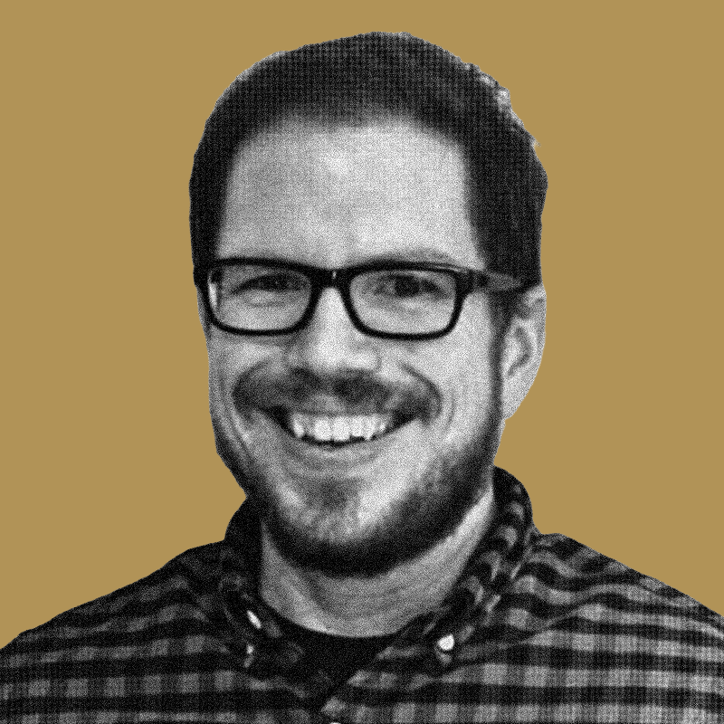 A smiling man with glasses, facial hair, and a checkered shirt against a plain background.