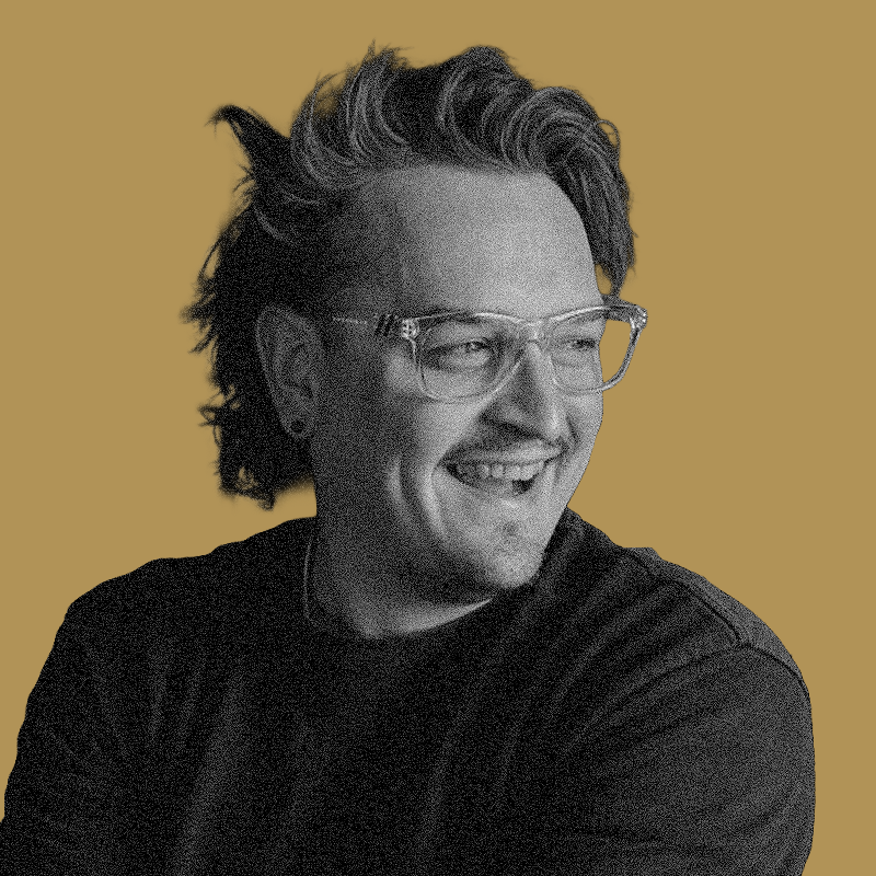 A smiling man with curly hair, glasses, and earrings wearing a dark shirt against a light brown background.