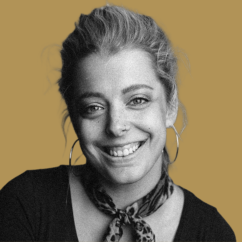 A smiling young woman with light skin, curly hair tied back, wearing hoop earrings, a patterned scarf, and a black top, set against a solid mustard background.
