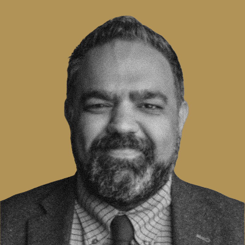 Black and white portrait of a man with a beard, wearing a suit and tie, with a neutral expression against a light brown background.