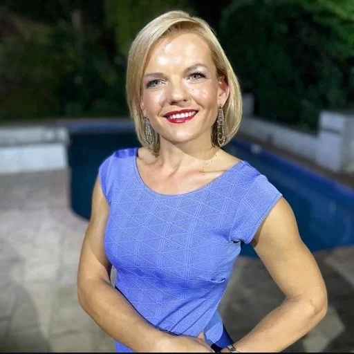 A woman with blonde hair wearing a blue dress, standing outdoors near a pool with trees in the background.