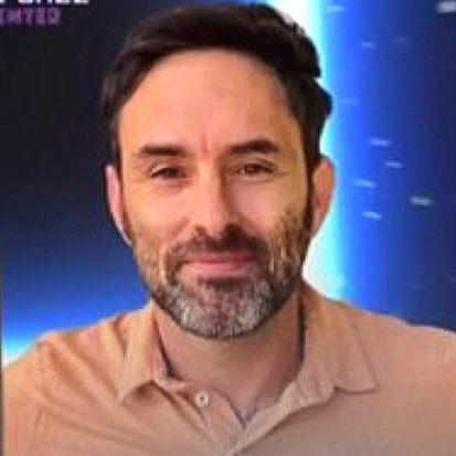 Close-up of a man with dark hair and a beard, wearing a light-colored collared shirt, smiling slightly.