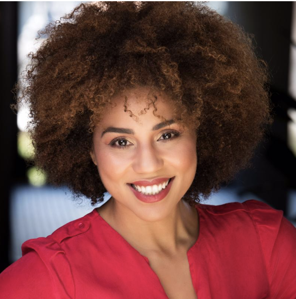 Smiling Joy Villa with curly brown hair wearing a red top, indoors with a blurred background.