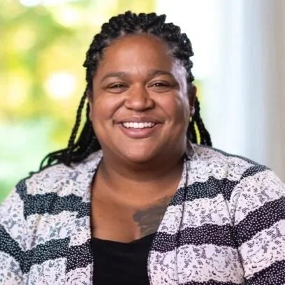 A smiling woman with braided hair, wearing a patterned blazer, sitting indoors with a window showing trees in the background.