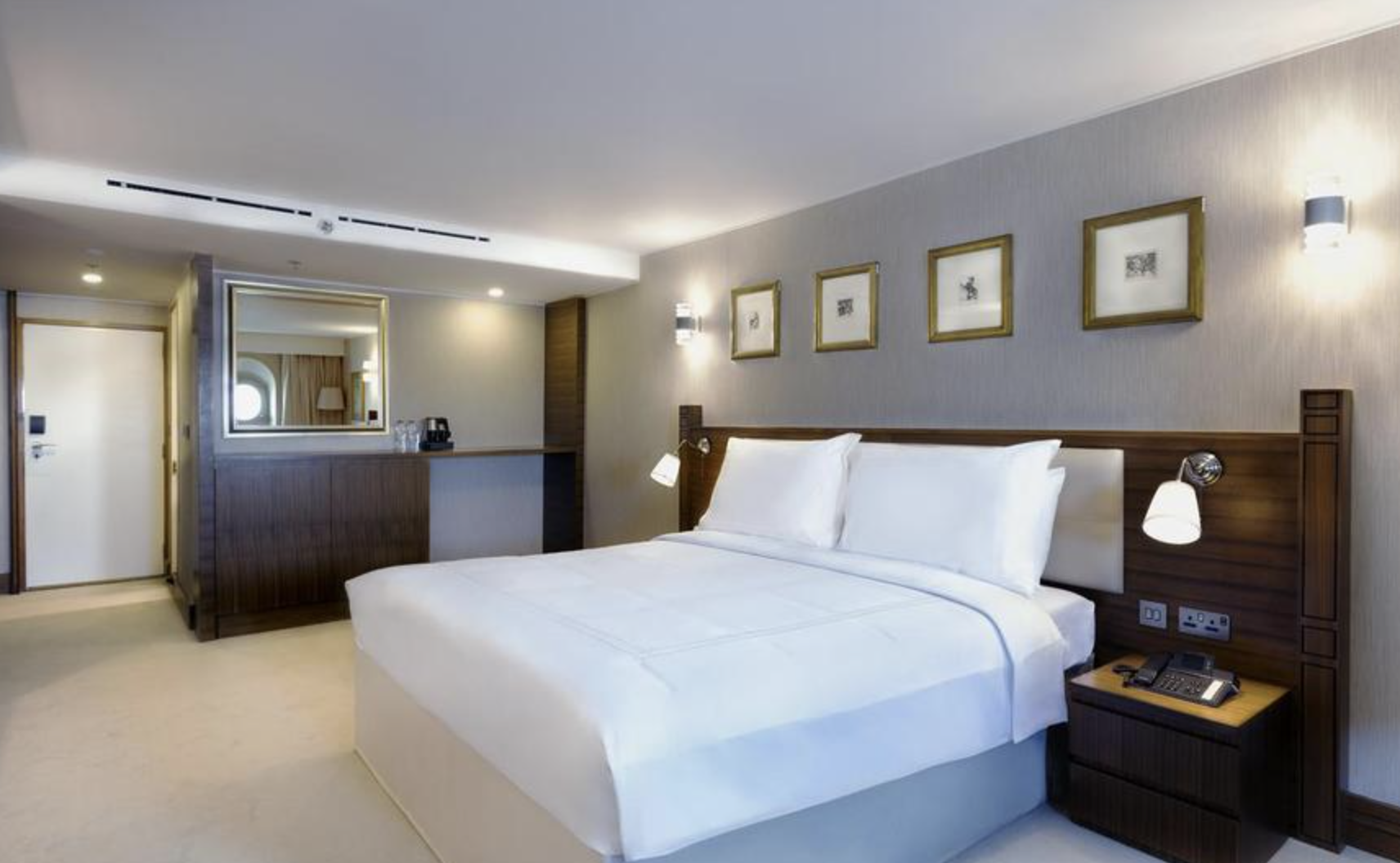 A hotel room with a neatly made bed, bedside lamps, artwork above the bed, and a small reception area with a mirror and coffee station.