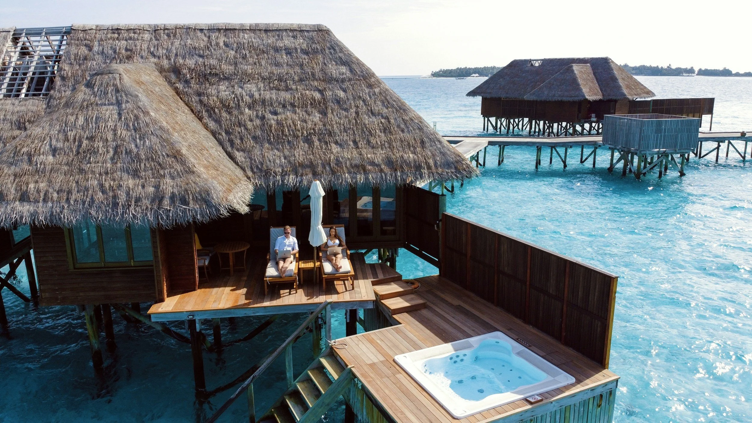 Overwater bungalow with thatched roof, featuring a hot tub on the wooden deck and two people relaxing on lounge chairs under a large umbrella. The bungalow is situated above clear blue water with another similar structure in the background.