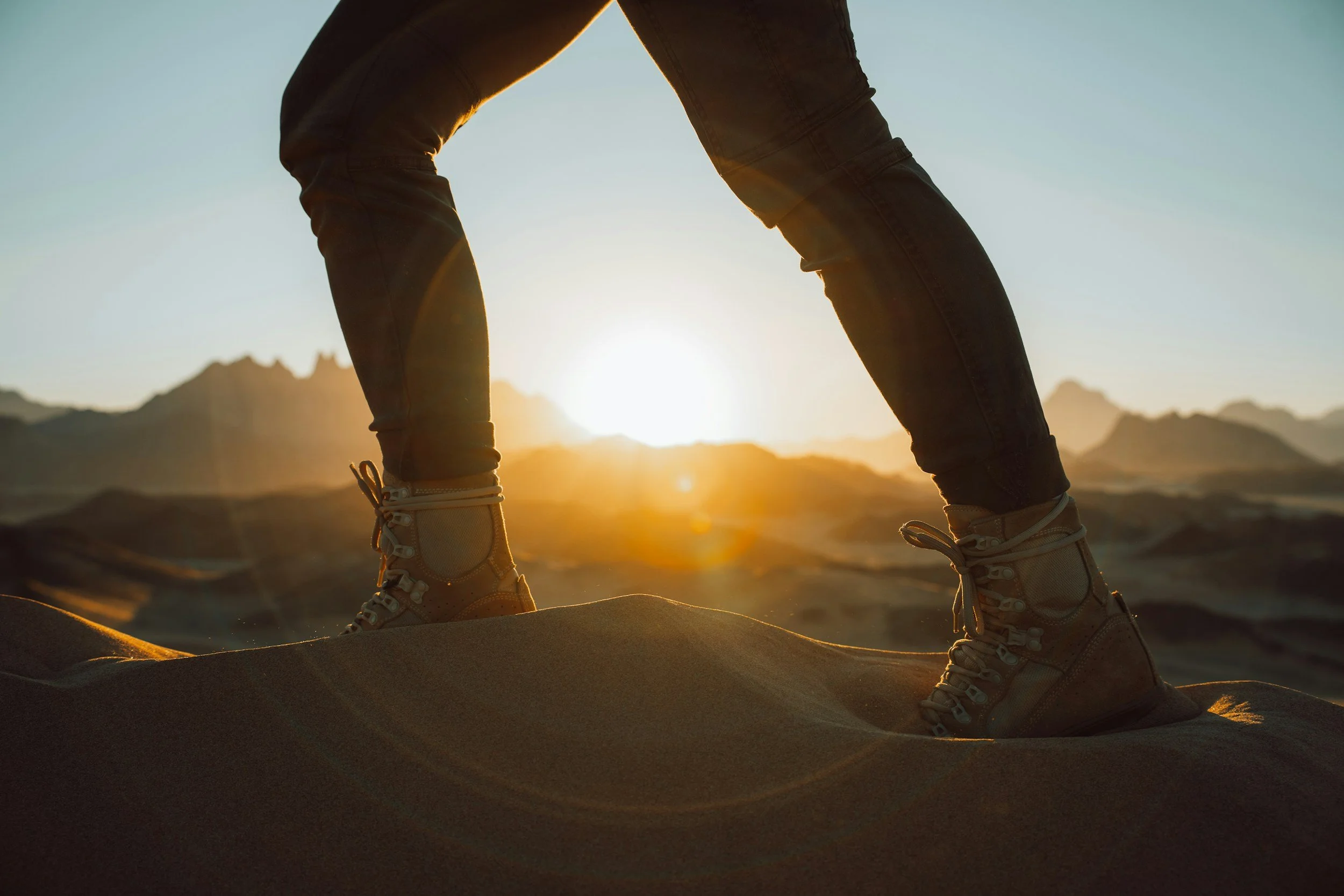 Person in hiking boots bending over on sandy terrain with mountains in the background during sunset.