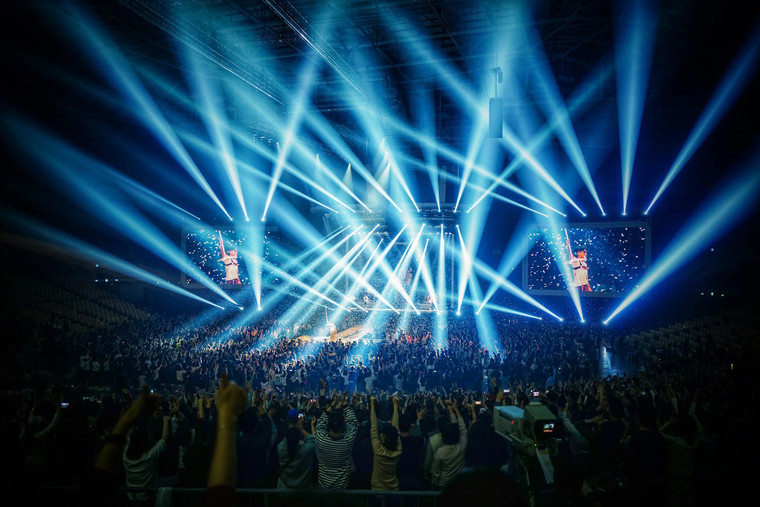 Crowd attending a concert with bright blue stage lights and large screens displaying performers.