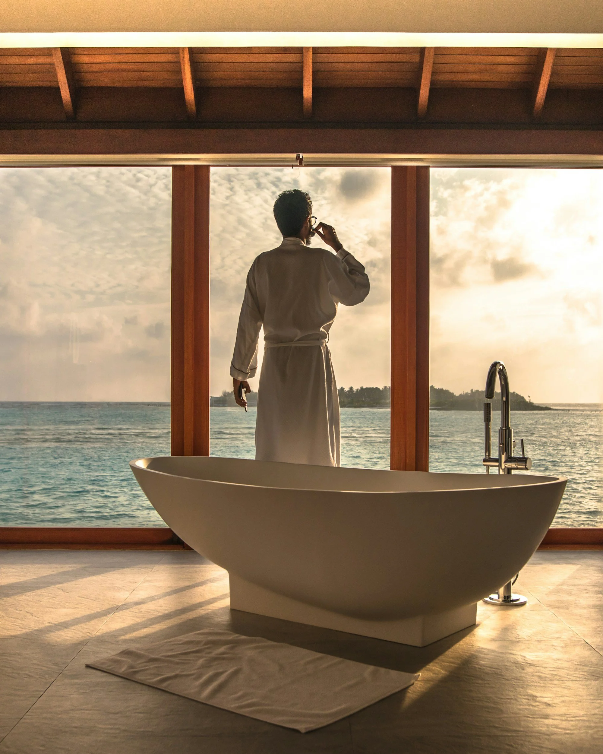 A man in a bathrobe looking out a large window at the ocean during sunset, with a bathtub in the foreground inside a room.