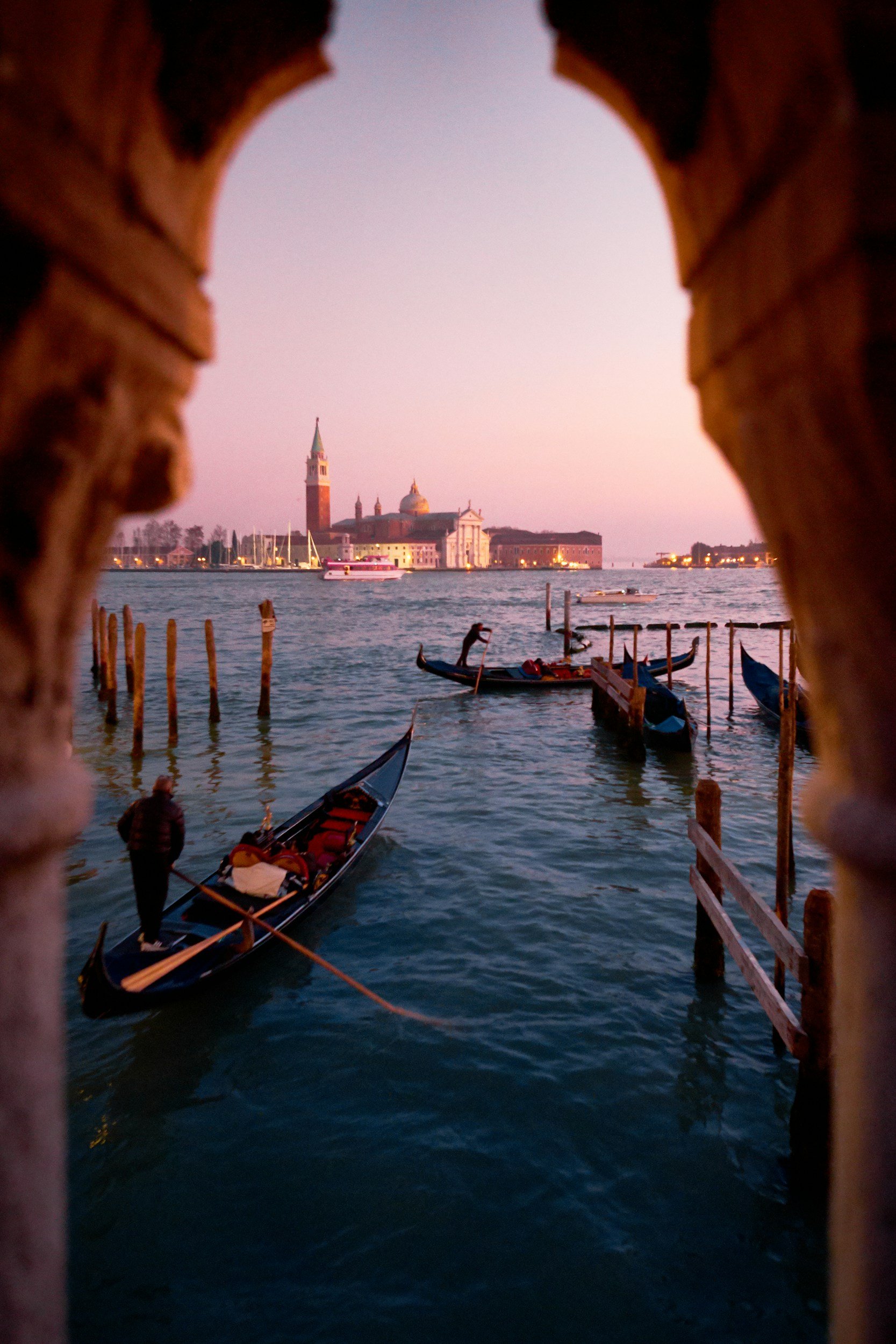 View of Venice, Italy, through a small opening, showing gondolas on the water and historic buildings, including a church with a tall bell tower, during sunset.