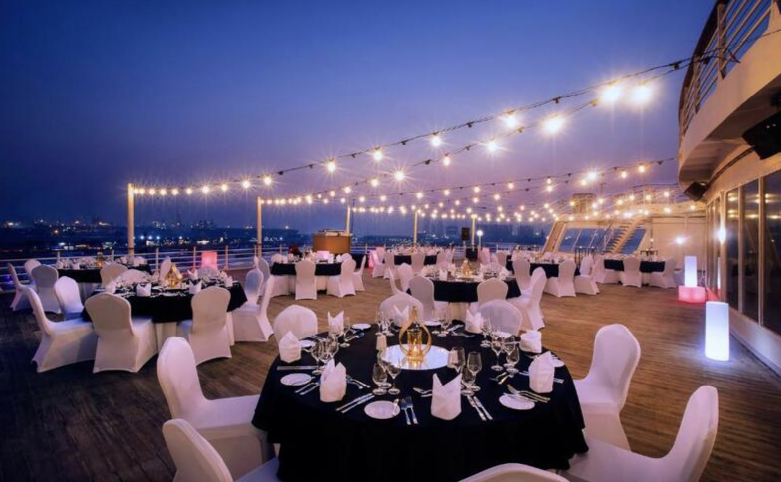 Outdoor dinner setup on a boat deck with round tables, white chairs, and string lights at dusk.