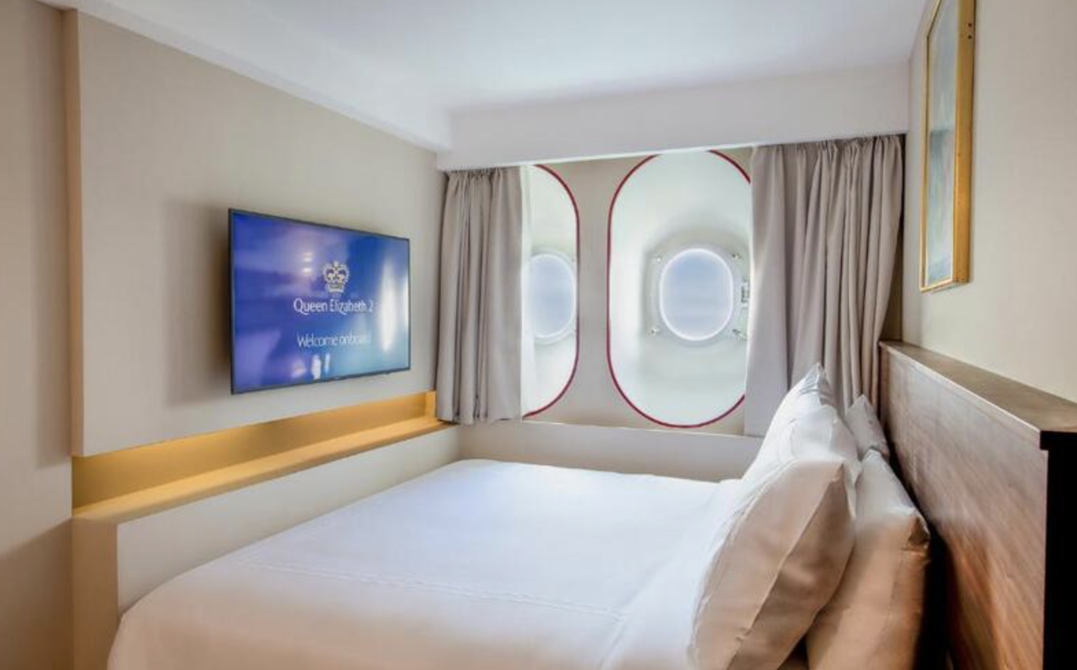 Hotel room with a bed, a wall-mounted TV displaying 'Queen Elizabeth' and 'Welcome onboard,' curtains, and windows designed to look like airplane portholes.