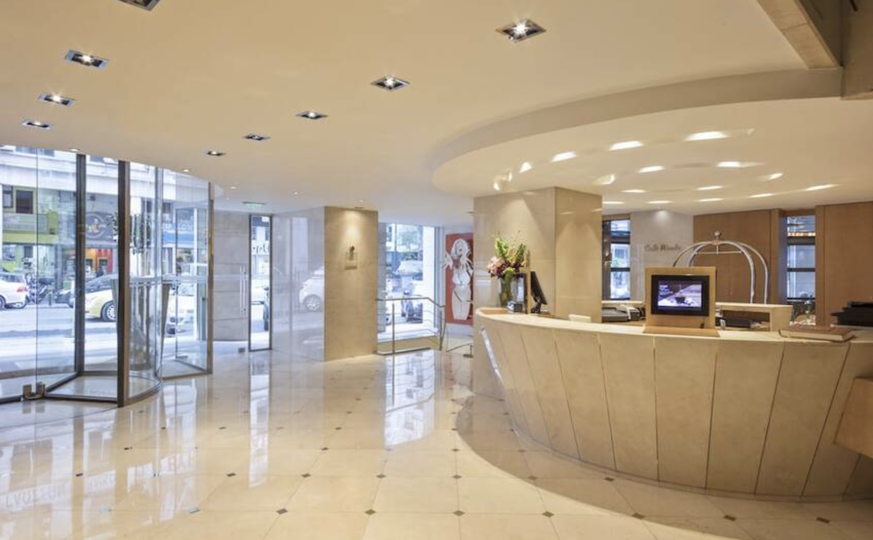Hotel lobby reception area with cream-colored curved desk, flowers, and a check-in computer, large glass windows showing street outside, polished marble floor, and soft ceiling lighting.