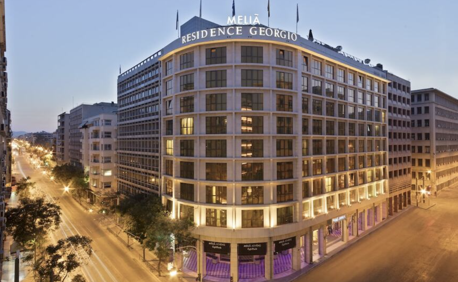 Night view of a modern multi-story hotel building with illuminated windows and signs, located on a city street with trees and streetlights.