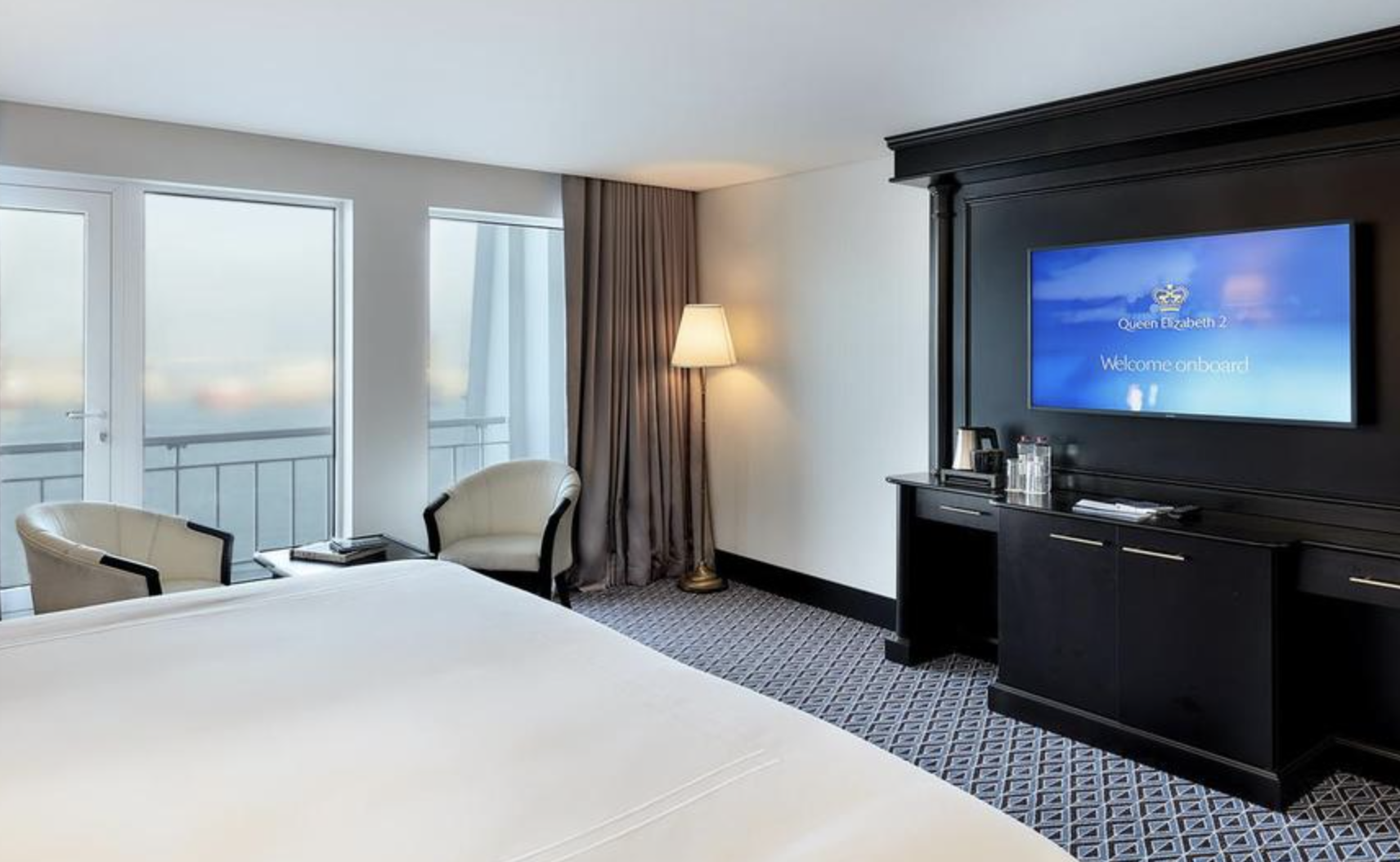 Hotel room with a bed, two chairs near large windows, a lamp, and a black entertainment center with a TV showing a welcome message for Queen Elizabeth 2.