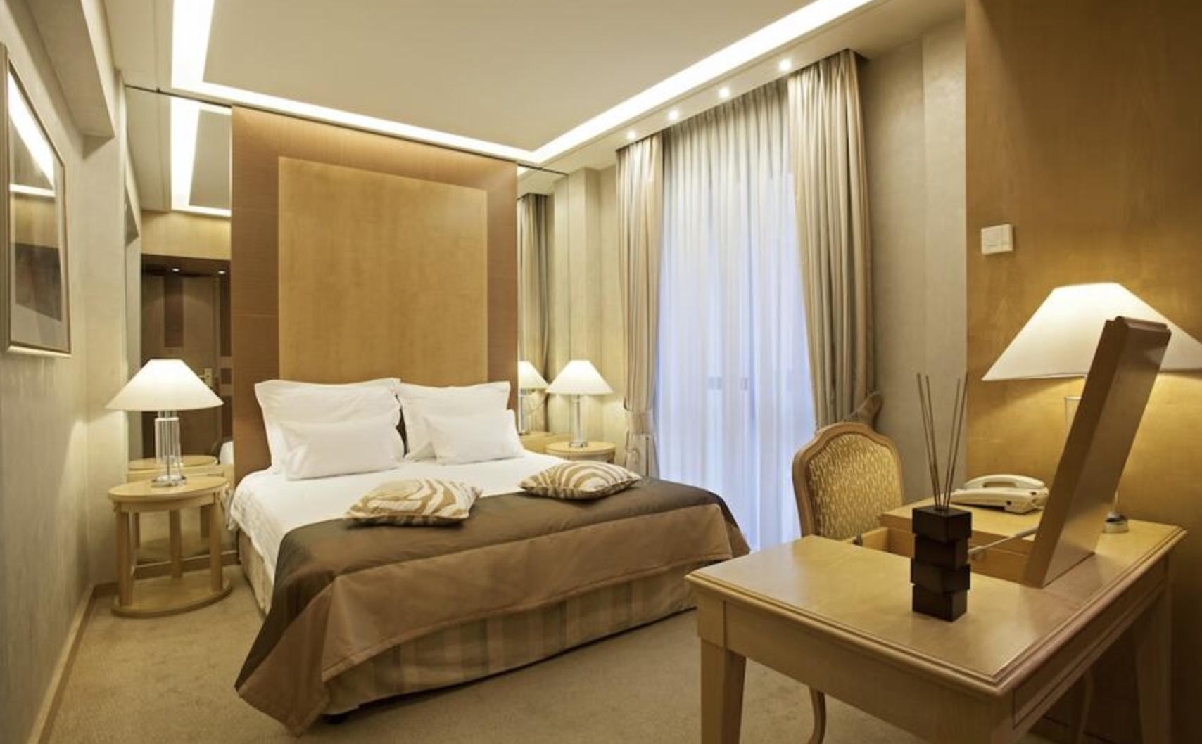 Hotel room with a bed, bedside lamps, a desk with a lamp, and a window with curtains.