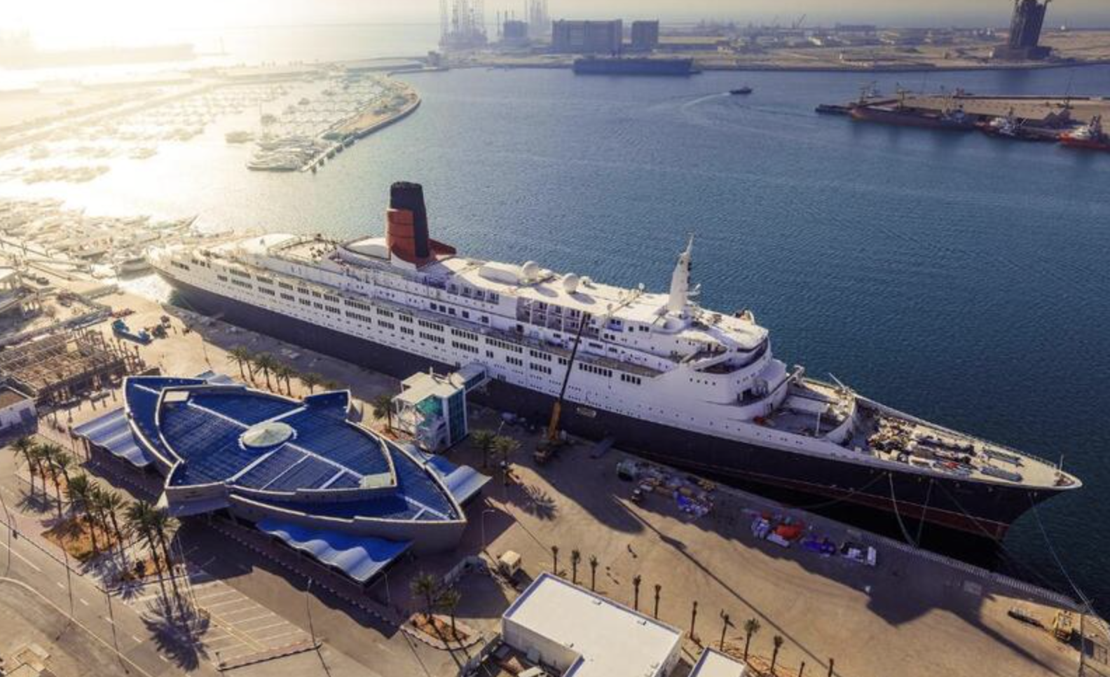 A cruise ship docked at a harbor with a modern building with a blue roof in the foreground, palm trees lining the streets, and other ships and buildings visible in the background.