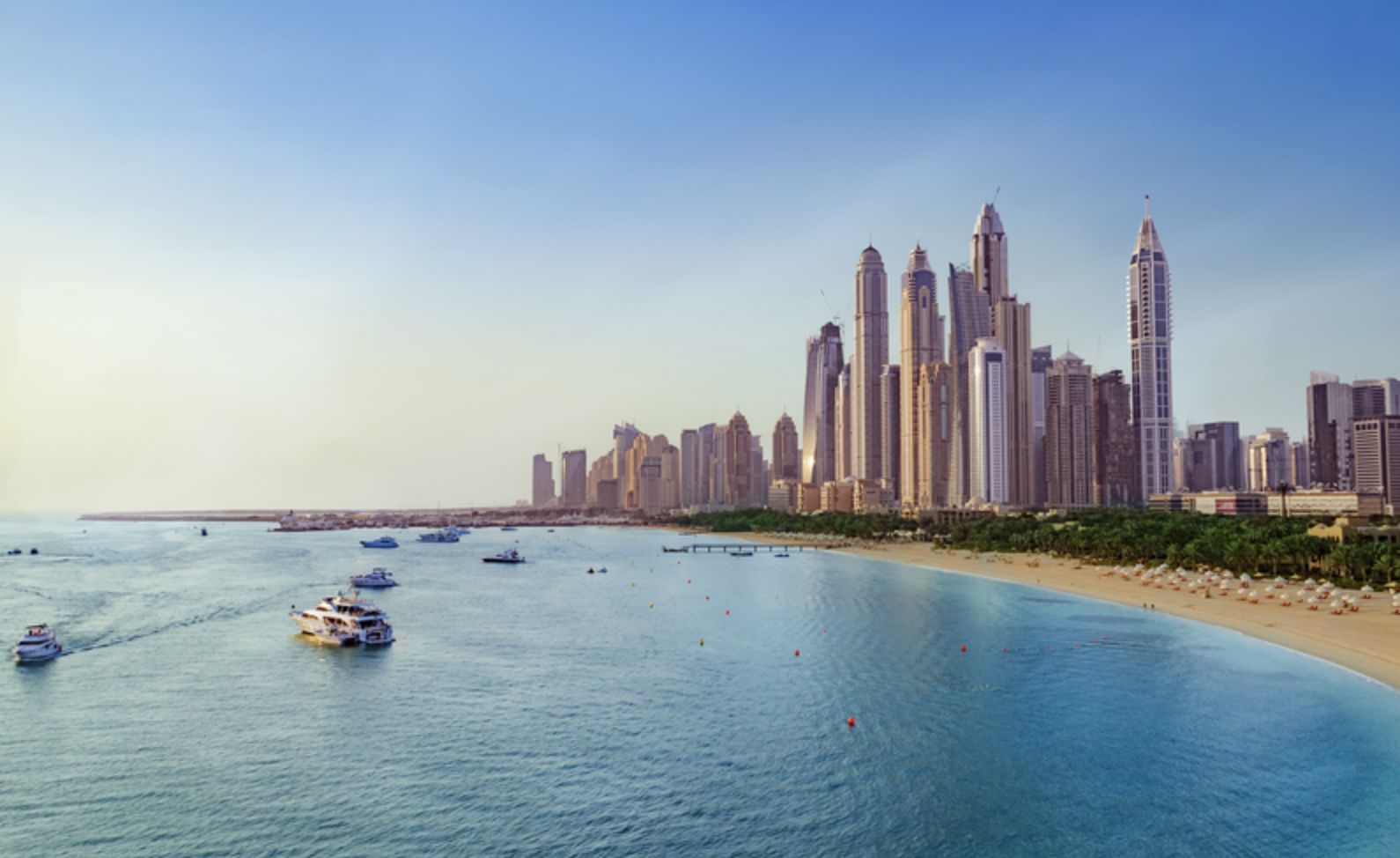 Skyline of Miami, Florida, with tall skyscrapers near the waterfront, several boats on calm water, and a sandy beach with umbrellas in the foreground.