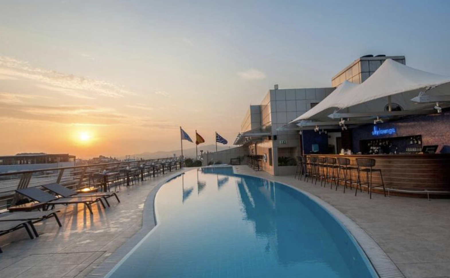 Rooftop pool area at sunset with lounge chairs and flags, adjacent to a bar with a sign for sky lounge.