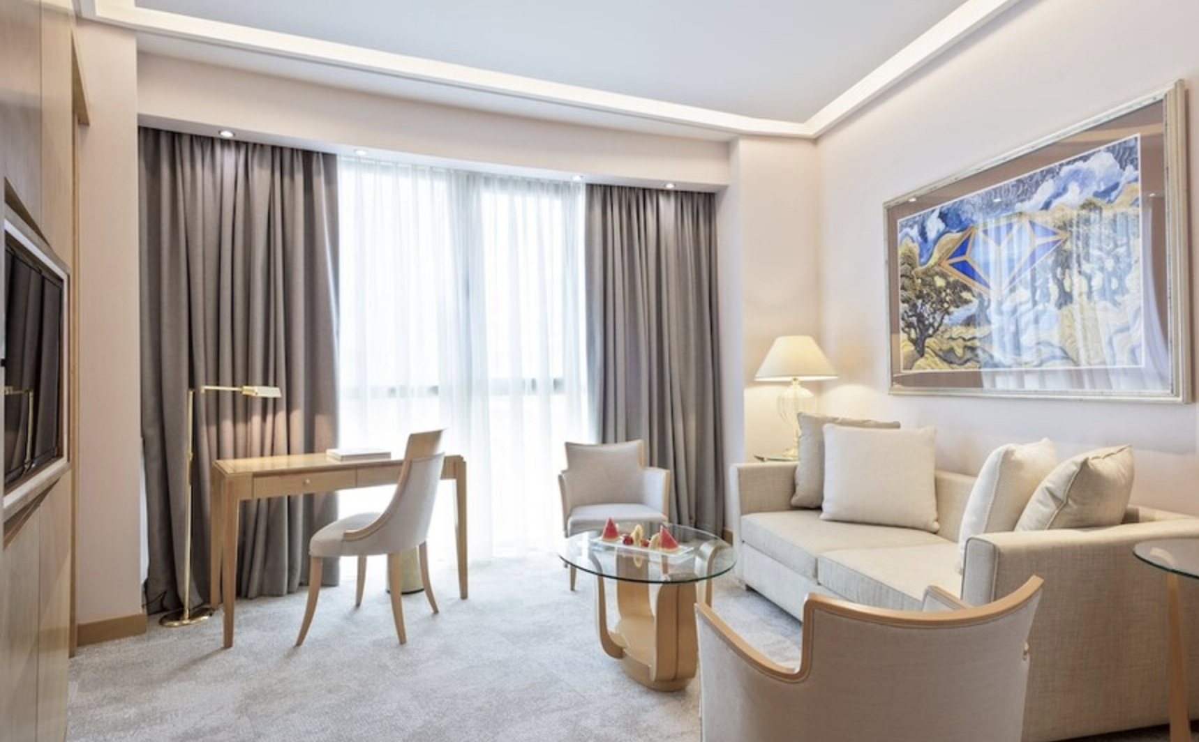 Elegant hotel living room with a cream-colored sofa, matching armchairs, glass coffee table, desk with chair, large window with gray curtains, wall art, and a floor lamp.