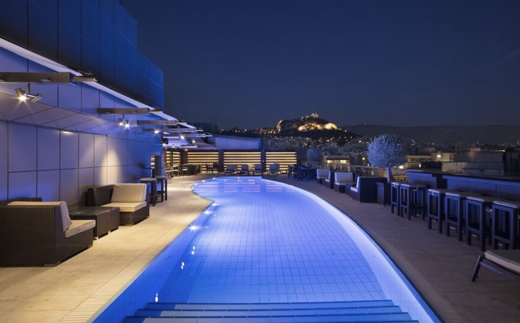 Luxury rooftop swimming pool at night with illuminated edges, surrounded by chairs and tables, overlooking a city with a hilltop fortress illuminated in the background.