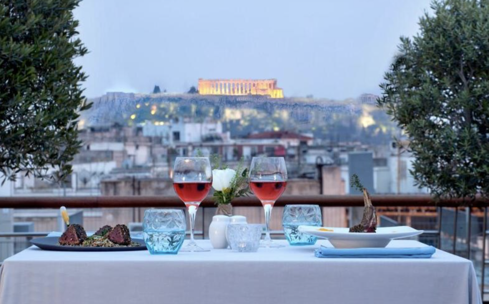 A dinner table with two glasses of rosé wine, plates of meat, a flower centerpiece, and a view of the Acropolis in Athens Greece in the background.