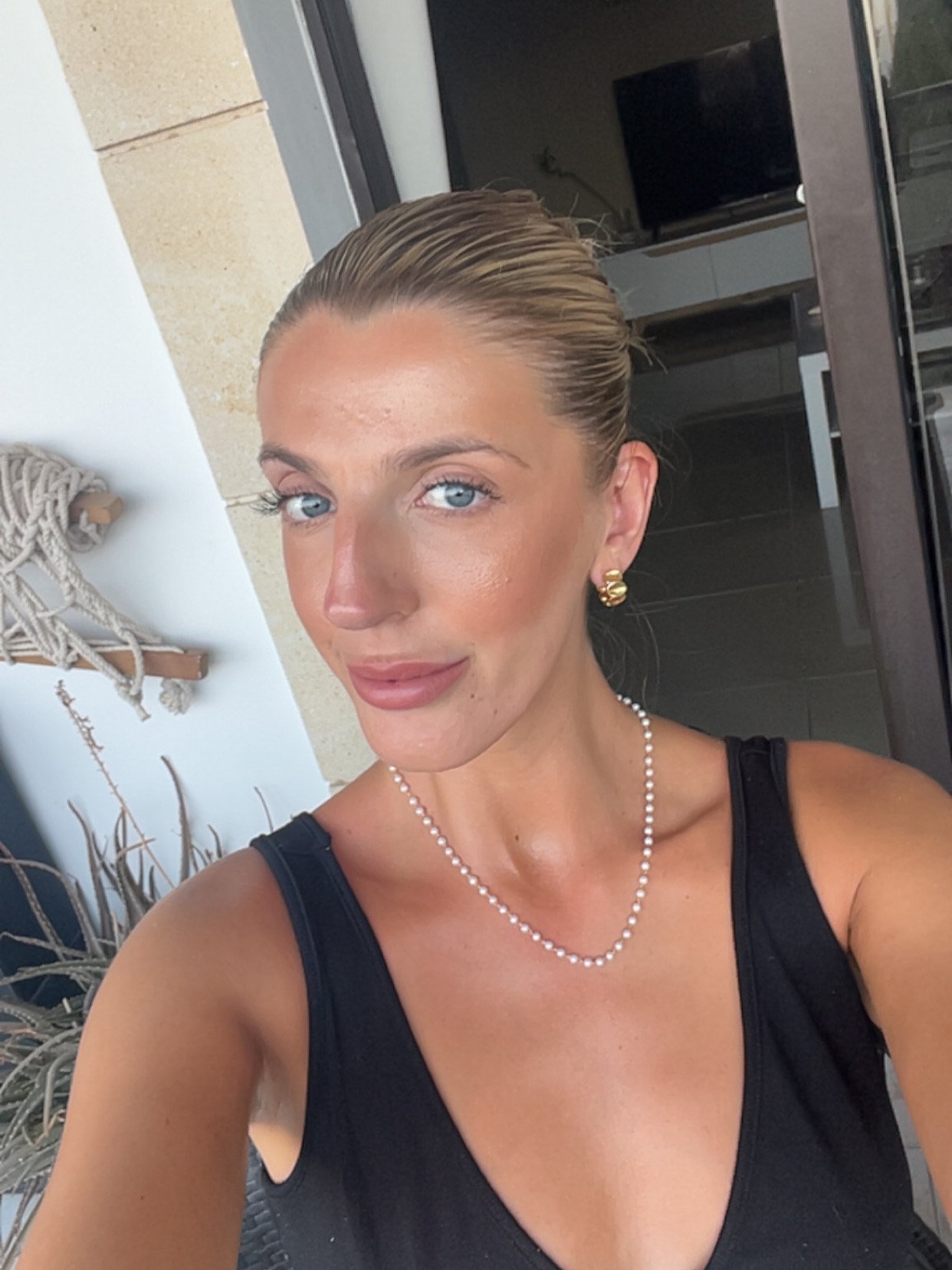 A woman with blonde hair tied back, wearing a black sleeveless top, pearl necklace, and gold earrings, taking a selfie outdoors near a building with a window and a TV in the background.
