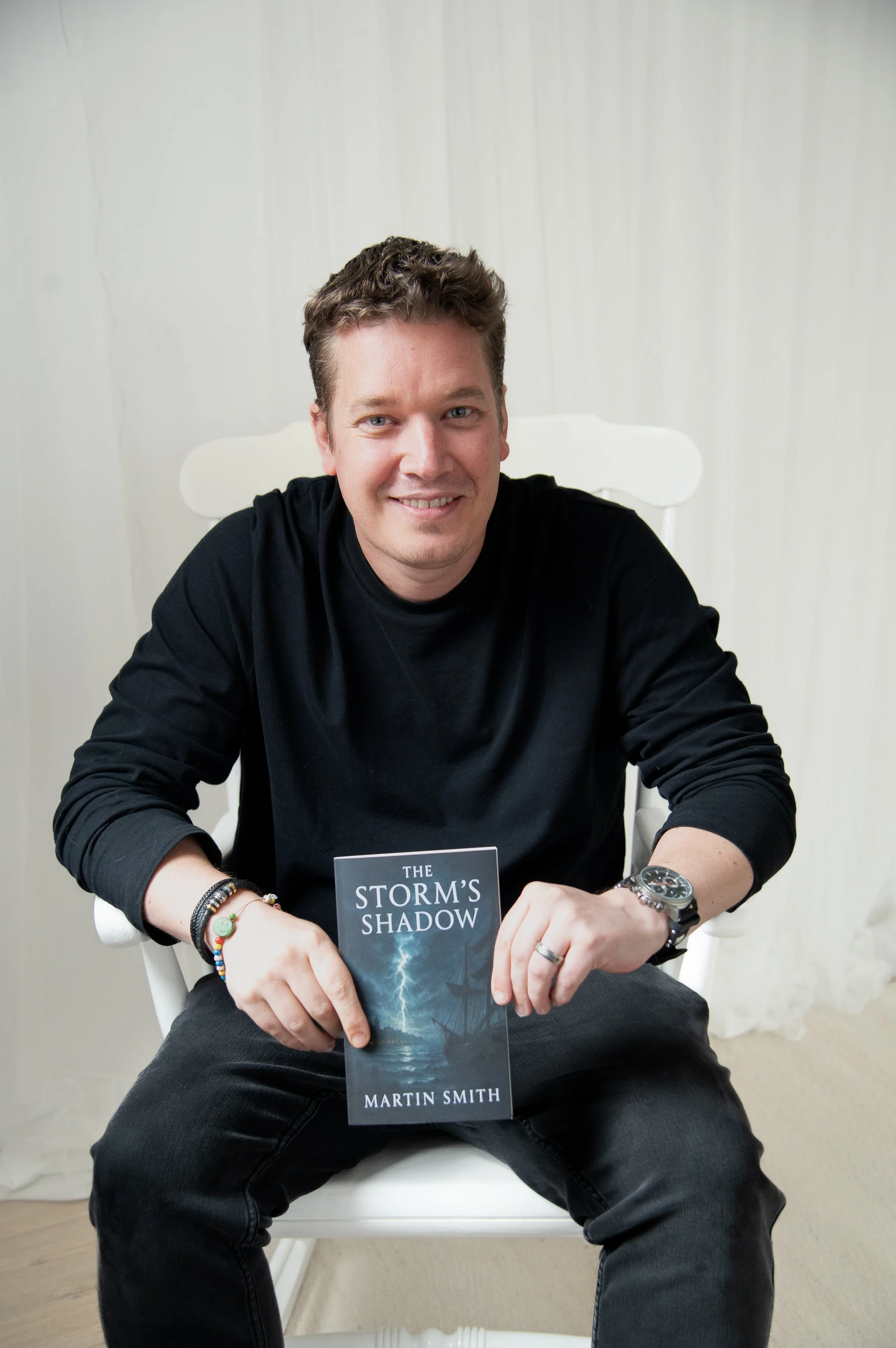 A young man with brown hair, wearing a black long-sleeve shirt, sitting on a white chair against a plain light-colored wall, holding a book titled 'The Storm's Shadow' by Martin Smith, and smiling at the camera.
