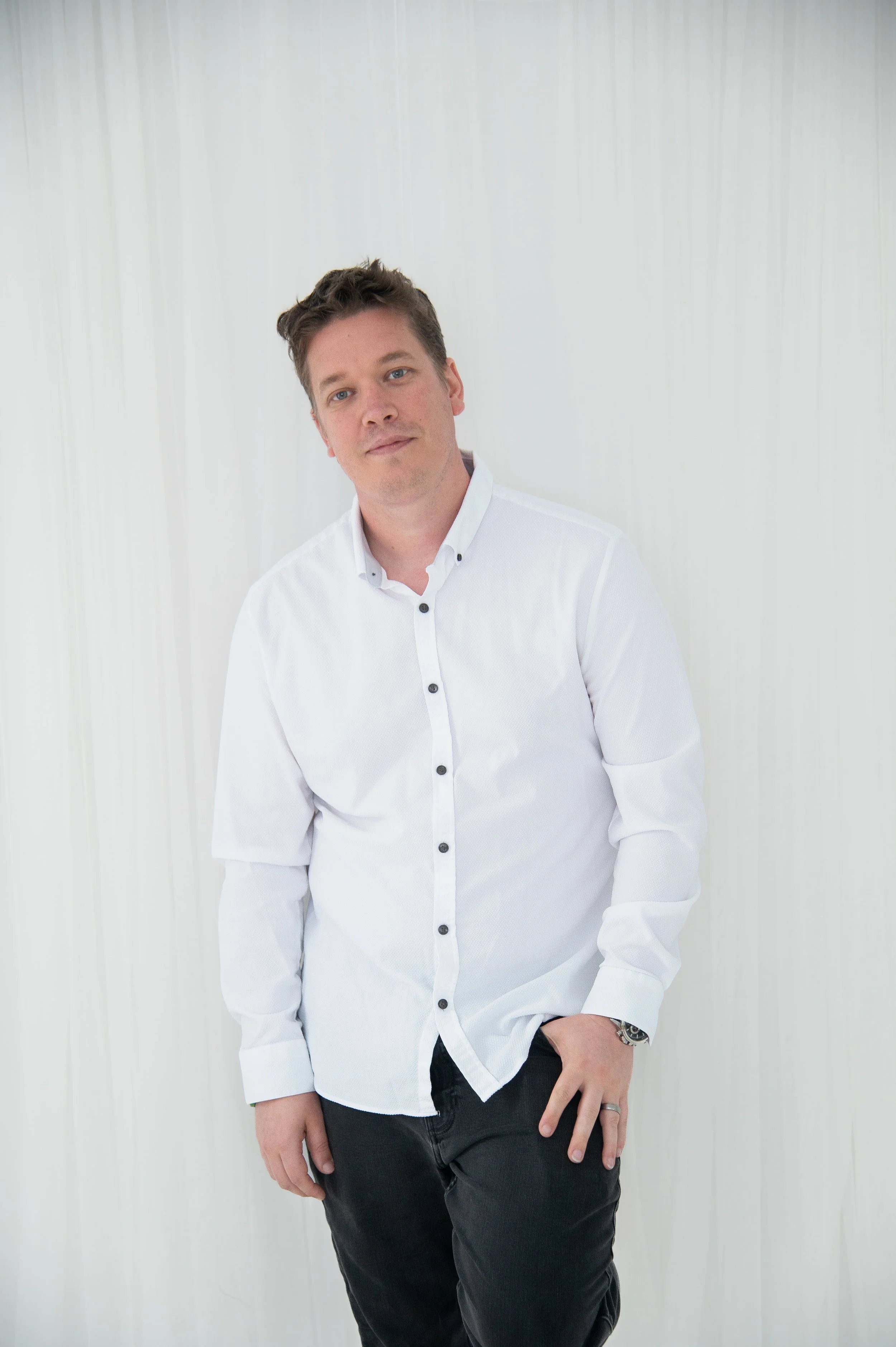 A young man with light skin and brown hair standing against a plain white wall, wearing a white button-up shirt and black pants.