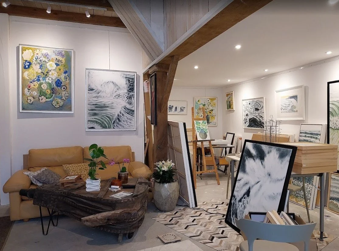 Une galerie d'art avec des peintures exposées sur les murs et en train d'être installées, un canapé beige, une table en bois rustique, des plantes, et un éclairage intérieur moderne.