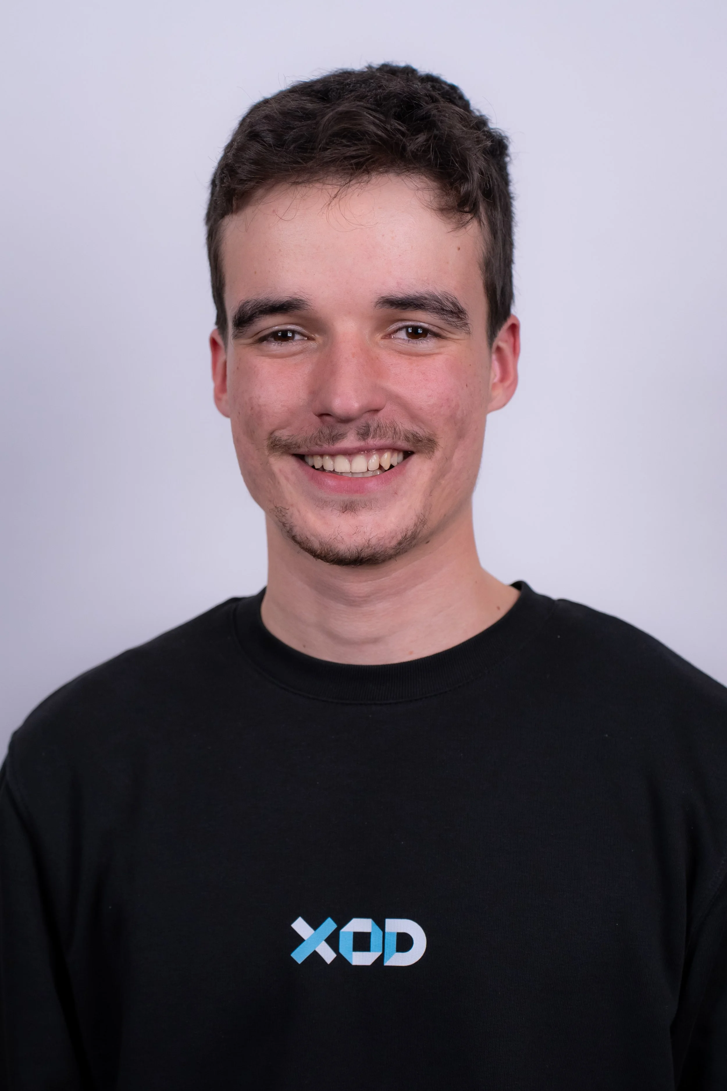 A woman with shoulder-length black hair, smiling, wearing a blue top, standing outdoors with green foliage in the background.