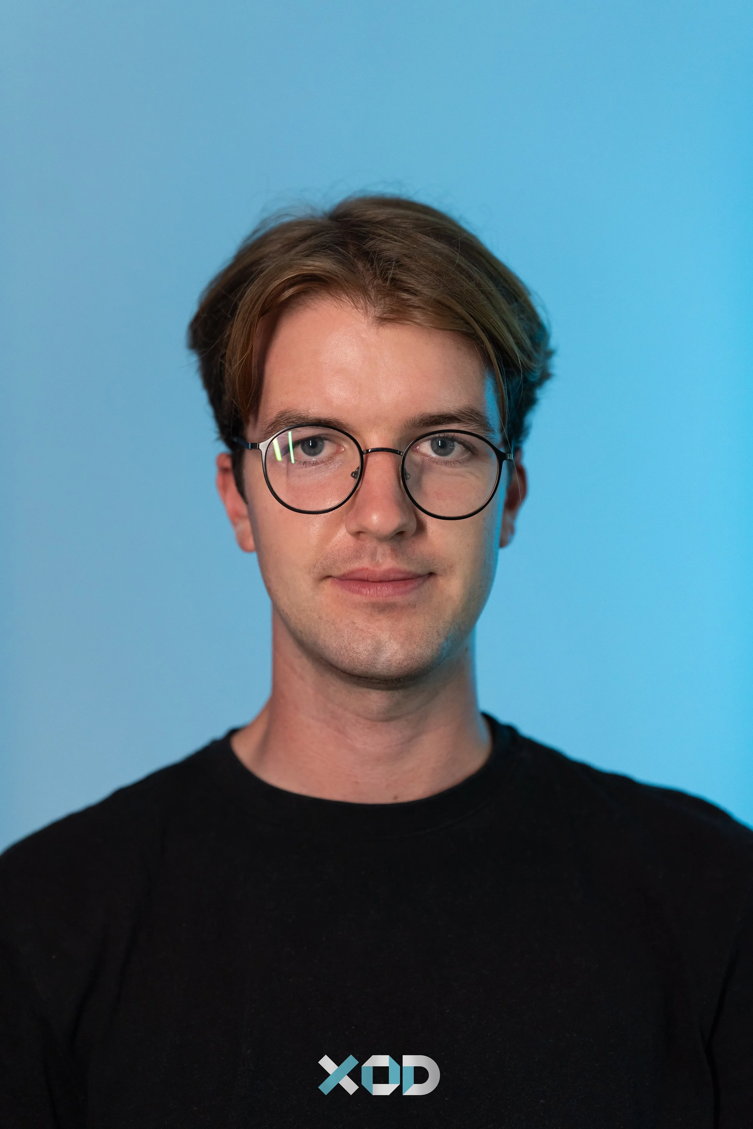 A man with curly dark hair, glasses, and a beard, wearing a dark green sweater, sitting indoors near a light-colored wall with a plant in the background.