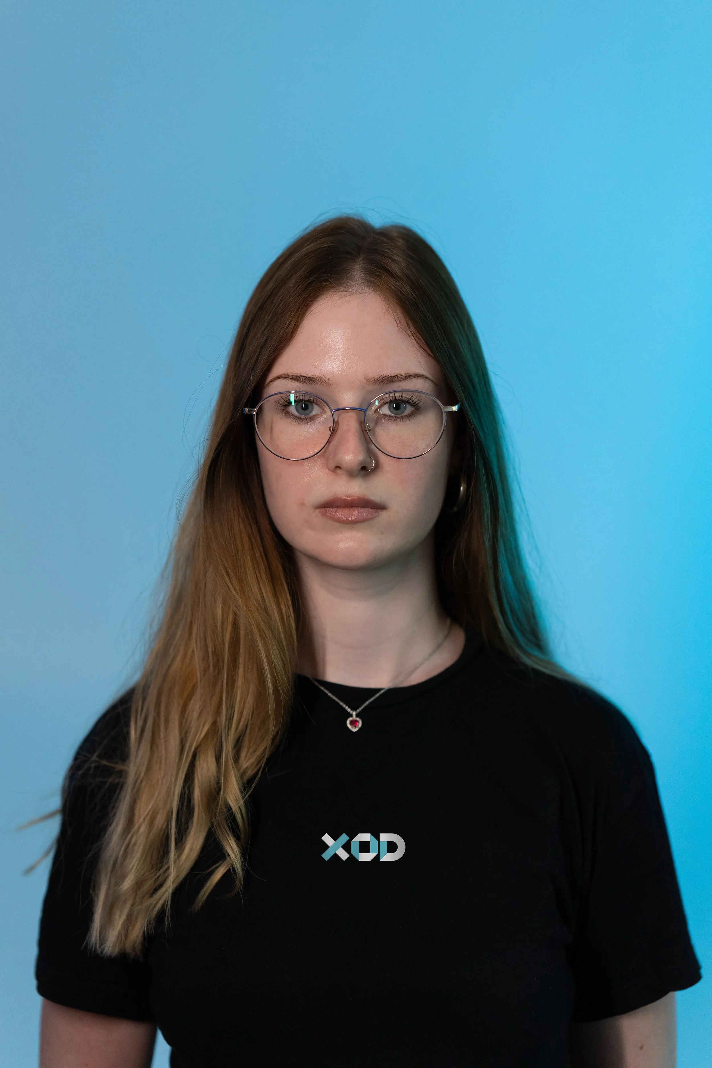 Portrait of a young woman with long, wavy blonde hair, wearing glasses, a black T-shirt with a logo, and a necklace with a red heart pendant, standing against a blue background.