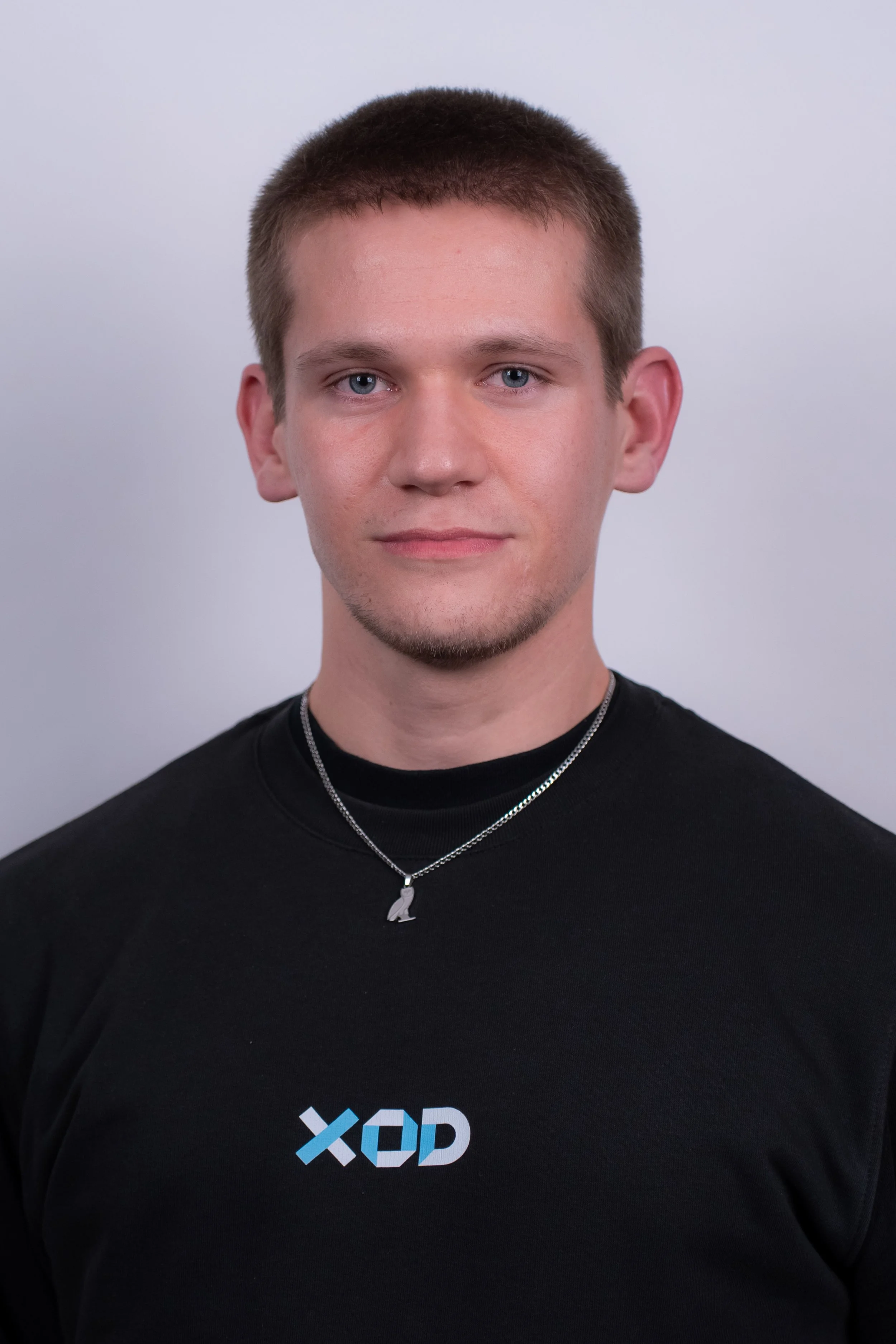 Young man with short hair and a goatee, wearing a black shirt with the letters 'XOD' on it and a silver necklace with a penguin pendant, standing against a blue background.