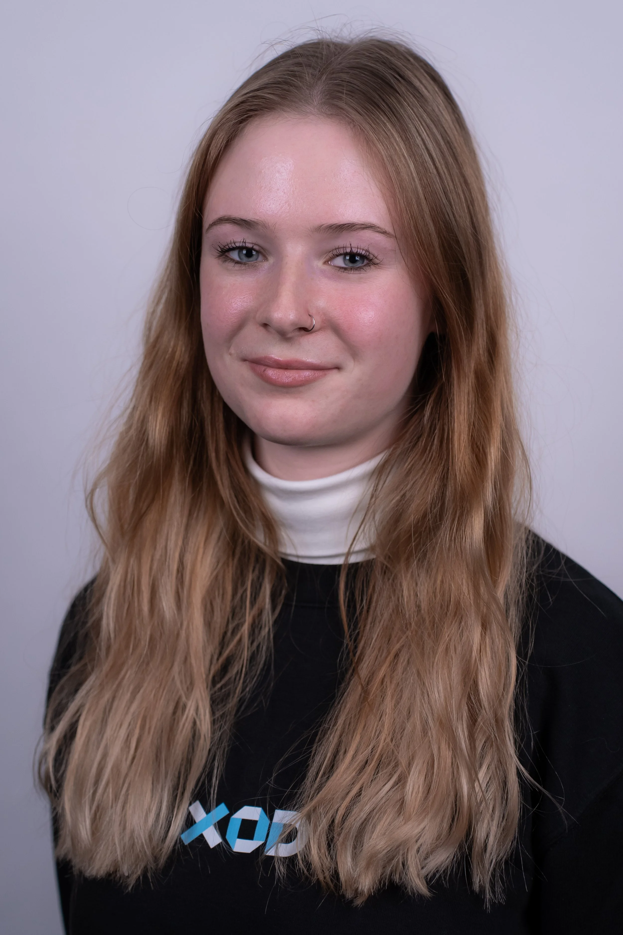 Portrait of a young woman with long, wavy blonde hair, wearing glasses, a black T-shirt with a logo, and a necklace with a red heart pendant, standing against a blue background.