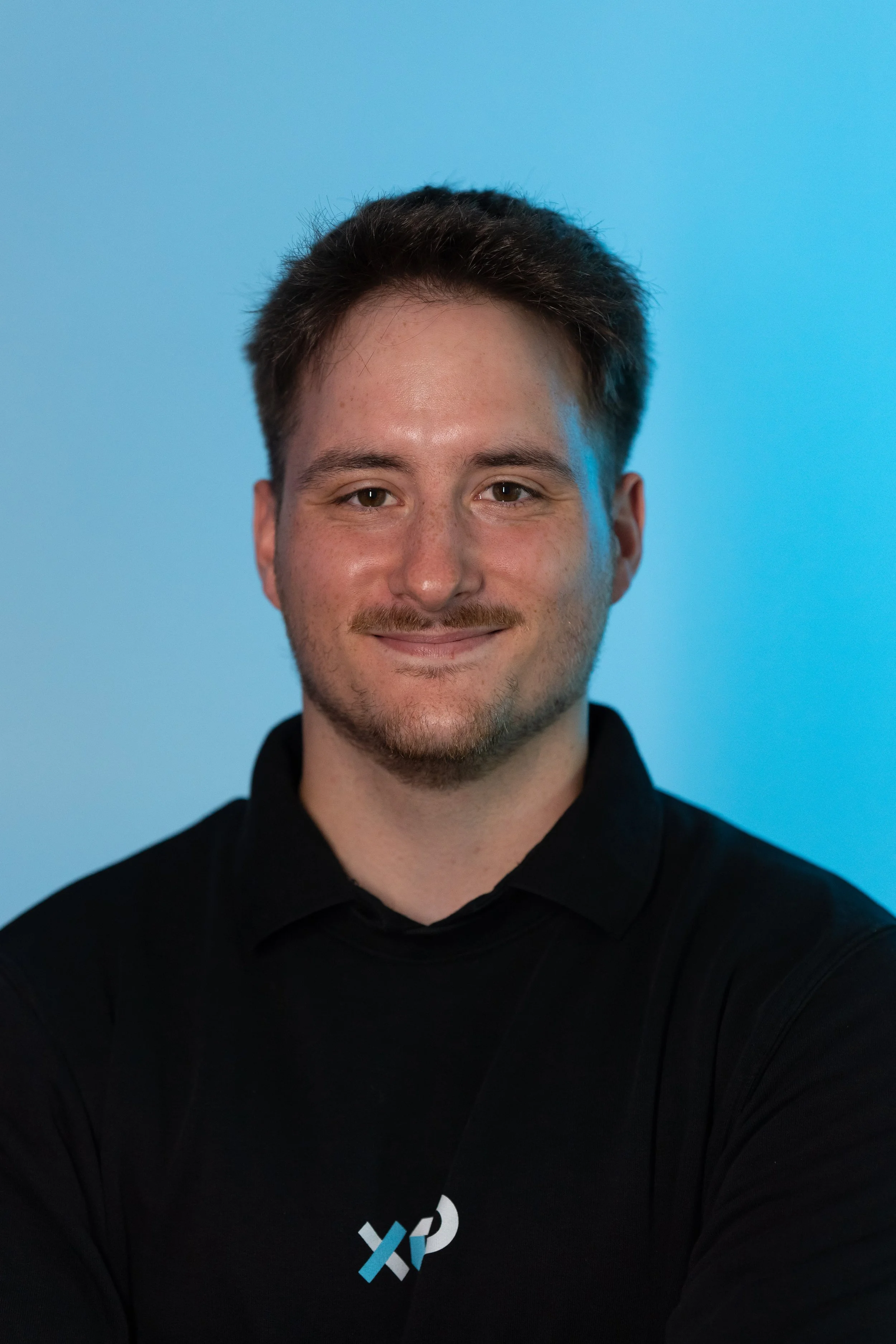 Headshot of a smiling young man with short brown hair, light facial hair, wearing a black shirt with a logo, against a blue background.